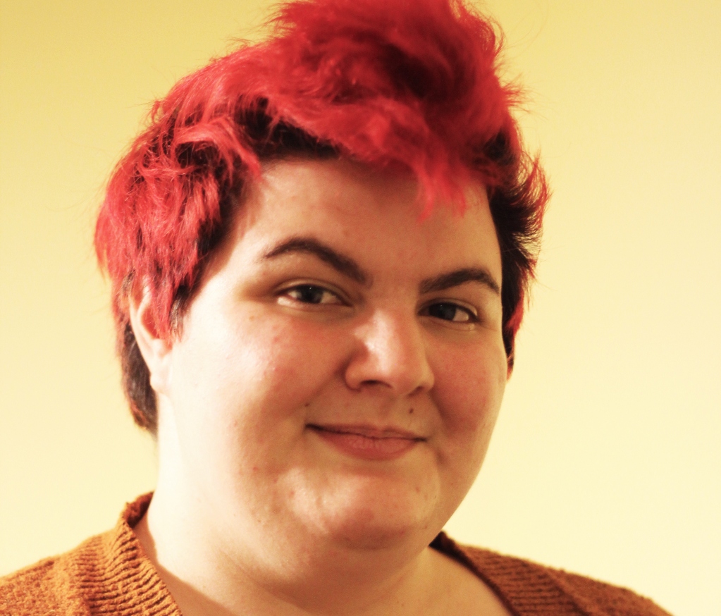 Sarah Kaufman headshot. They are a light skinned person with red hair and brown eyes. They are wearing an orange cardigan and smiling at the camera.